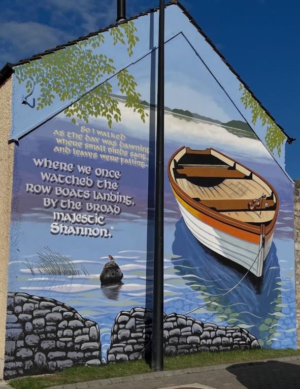 Mural at Emmet Place, Nenagh, by Neil O'Dwyer: a wooden rowing boat moored on still water beneath a tree, with lettered lines from Shane MacGowan's song The Broad Majestic Shannon painted alongside it