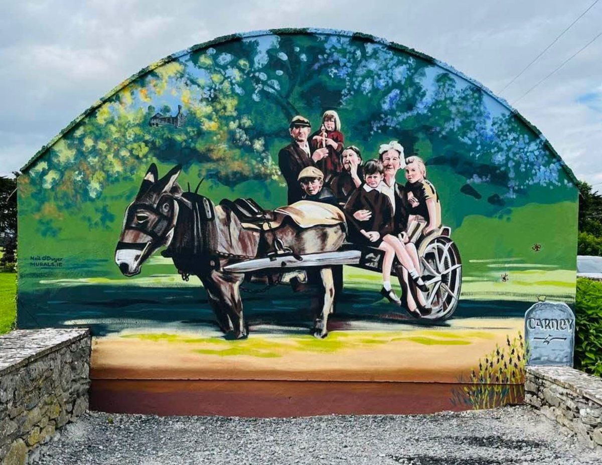 Mural at the Carney signpost, Co. Tipperary, by Neil O'Dwyer: a colour painting of an old photograph of Shane MacGowan as a boy in a cap driving a donkey and cart, with his sister Siobhán, their uncle and neighbours aboard